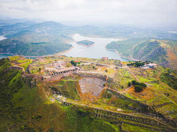 Ancient amphitheater nestled within sprawling ruins sits atop a verdant hill, overlooking a distant, serene lake and the surrounding lush, hilly landscape under a cloudy sky.